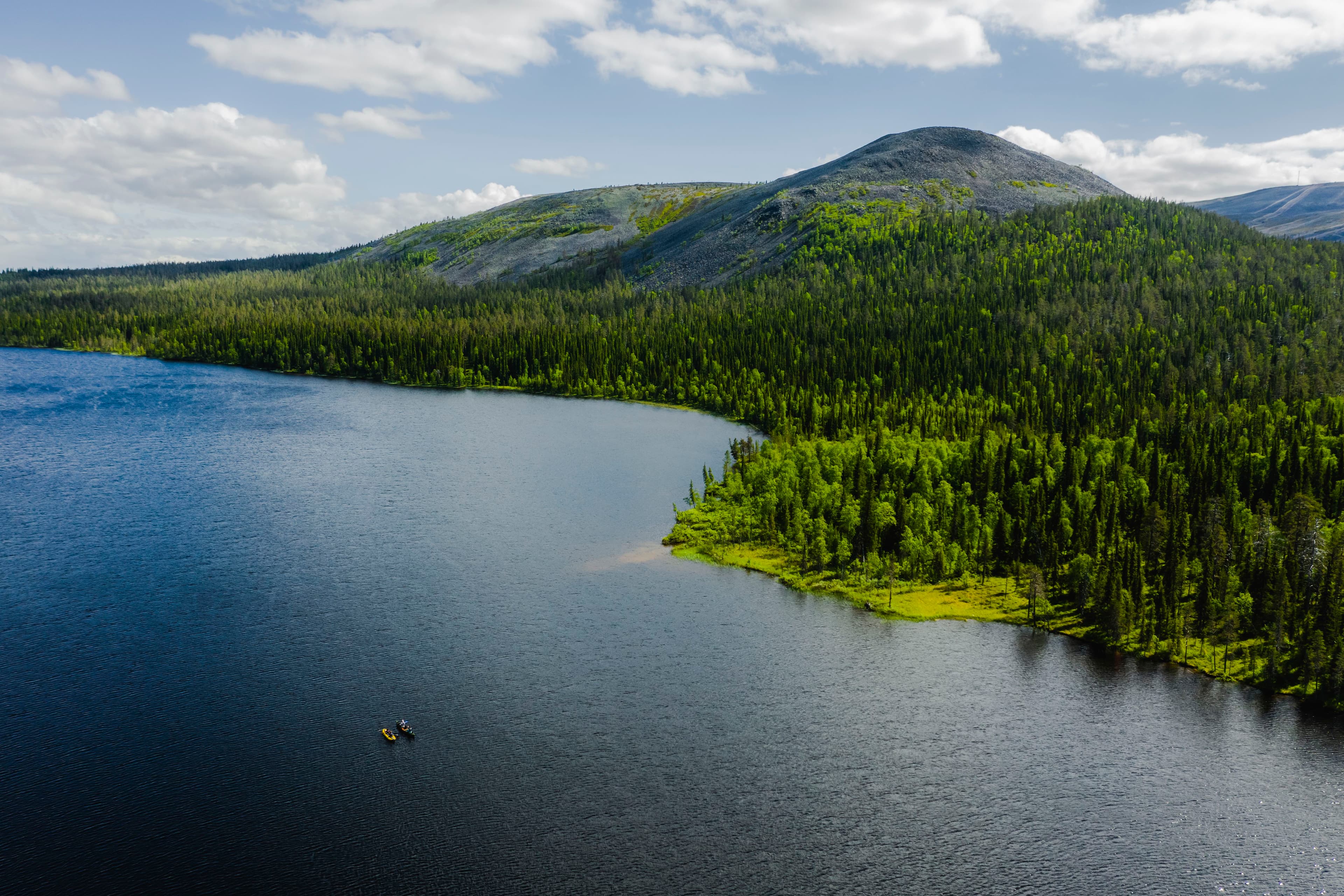 lmakuva rauhallisesta järvestä, jota reunustavat vehreät metsät ja kukkulat Ylläkseltä. Veden pinnalla pieni vene, taivas osittain pilvinen.
