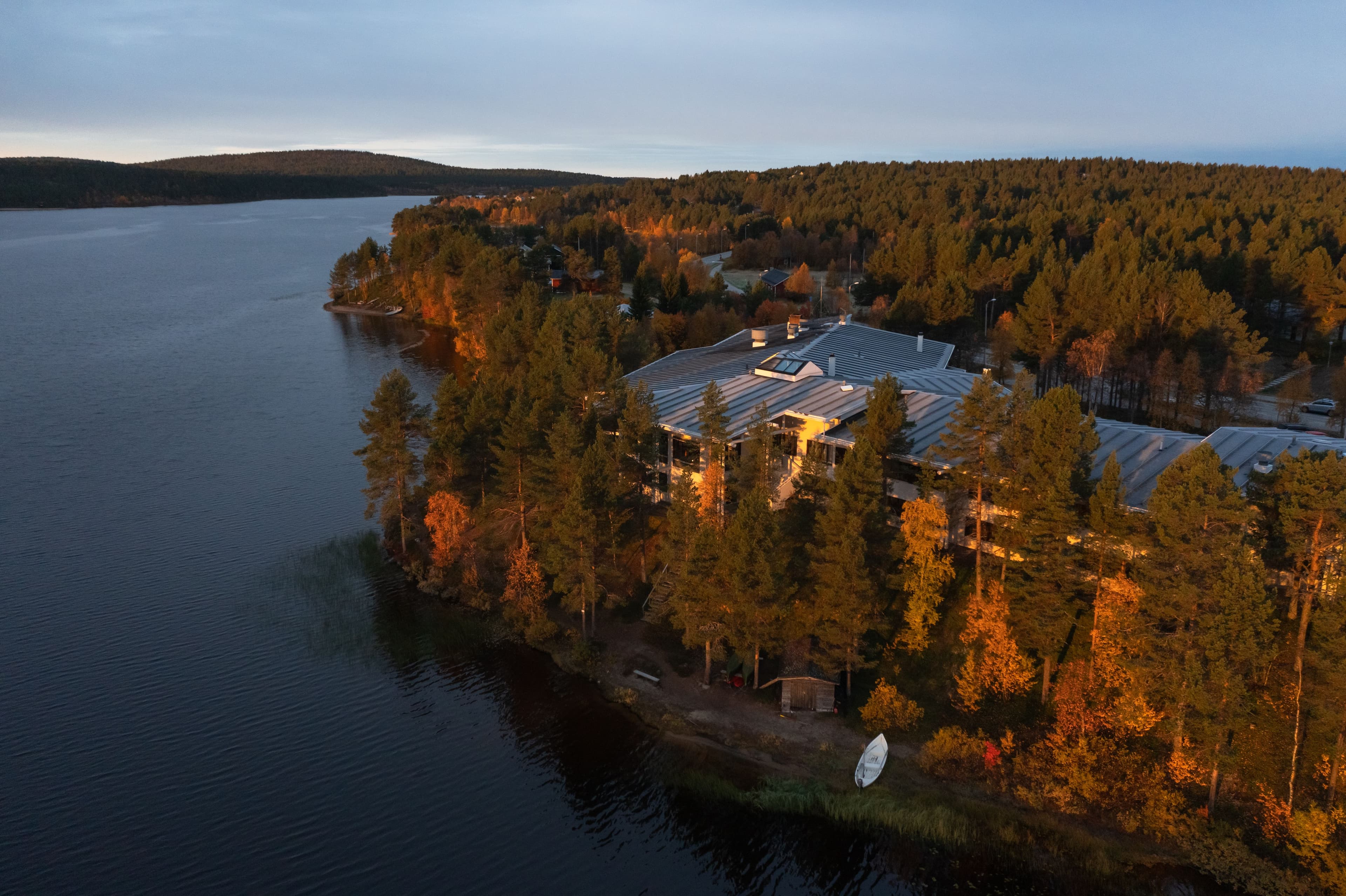 Ilmakuva Lapland Hotels Hetasta järven rannalla, ruskan värittämien metsien keskellä. Tyyni vesi heijastaa maiseman.