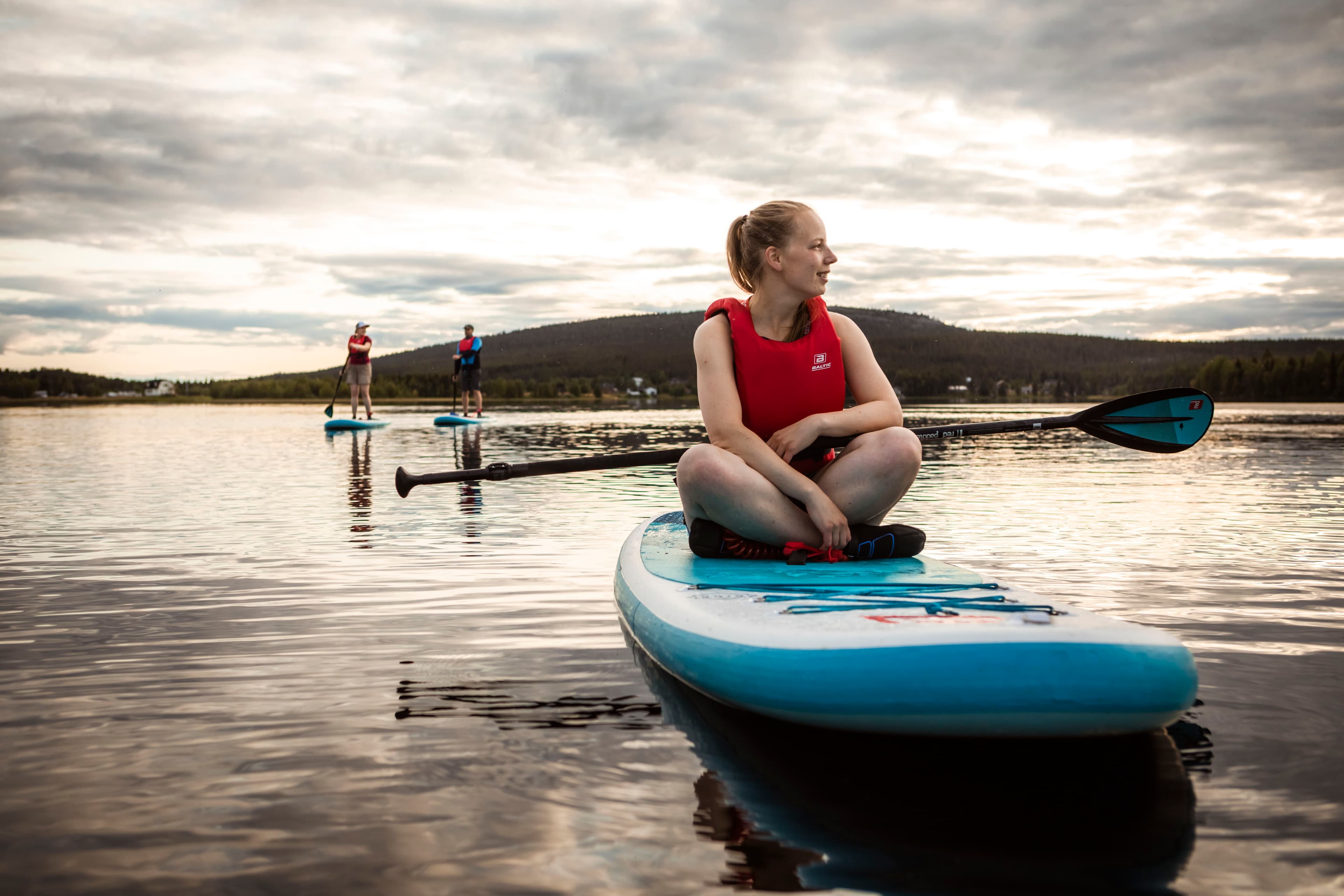 Nainen istuu suppilaudalla ja hymyilee. Taustalla kaksi ihmistä suppailee tyynellä järvellä pilvisen taivaan alla.